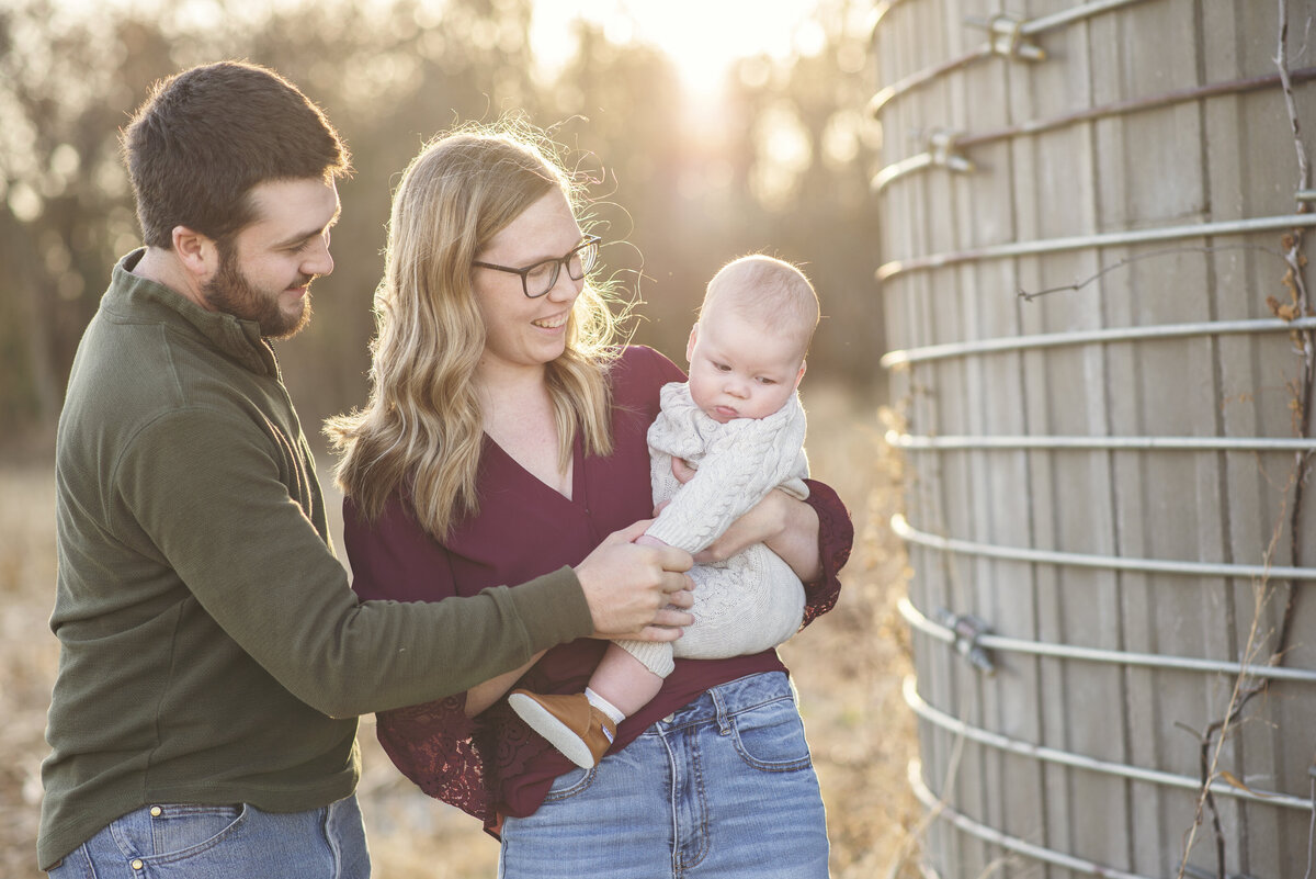 Parents standing near a silo, smiling as they help their baby boy stand during an outdoor family photo session.