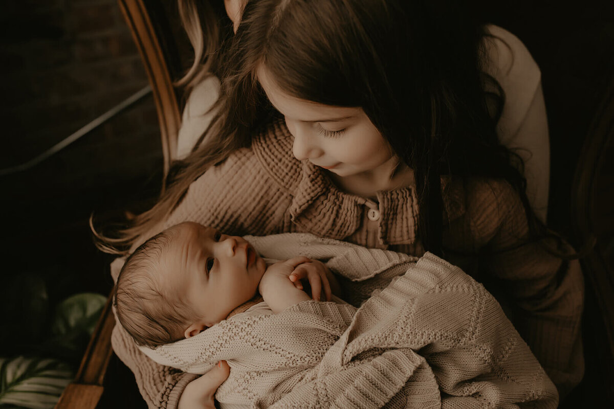 A new big sister cradlers her baby brother in her arms while they stare at each other