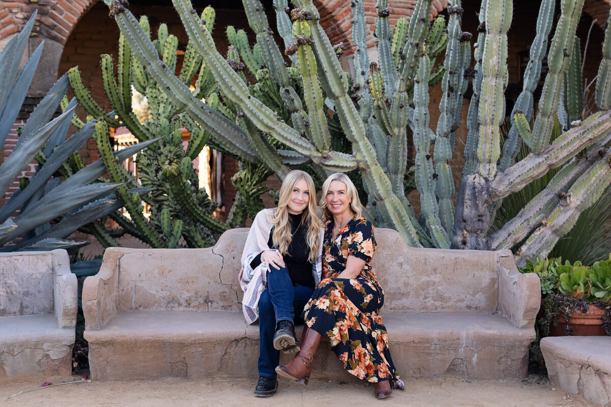 A distanced photo of a blonde mother wearing an orange and navy floral dress sitting on a bench next to her daughter with long, blonde hair, wearing blue jeans, a black turtleneck, and a grey and beige cardigan, both smiling at the camera with giant cactus plants behind them.