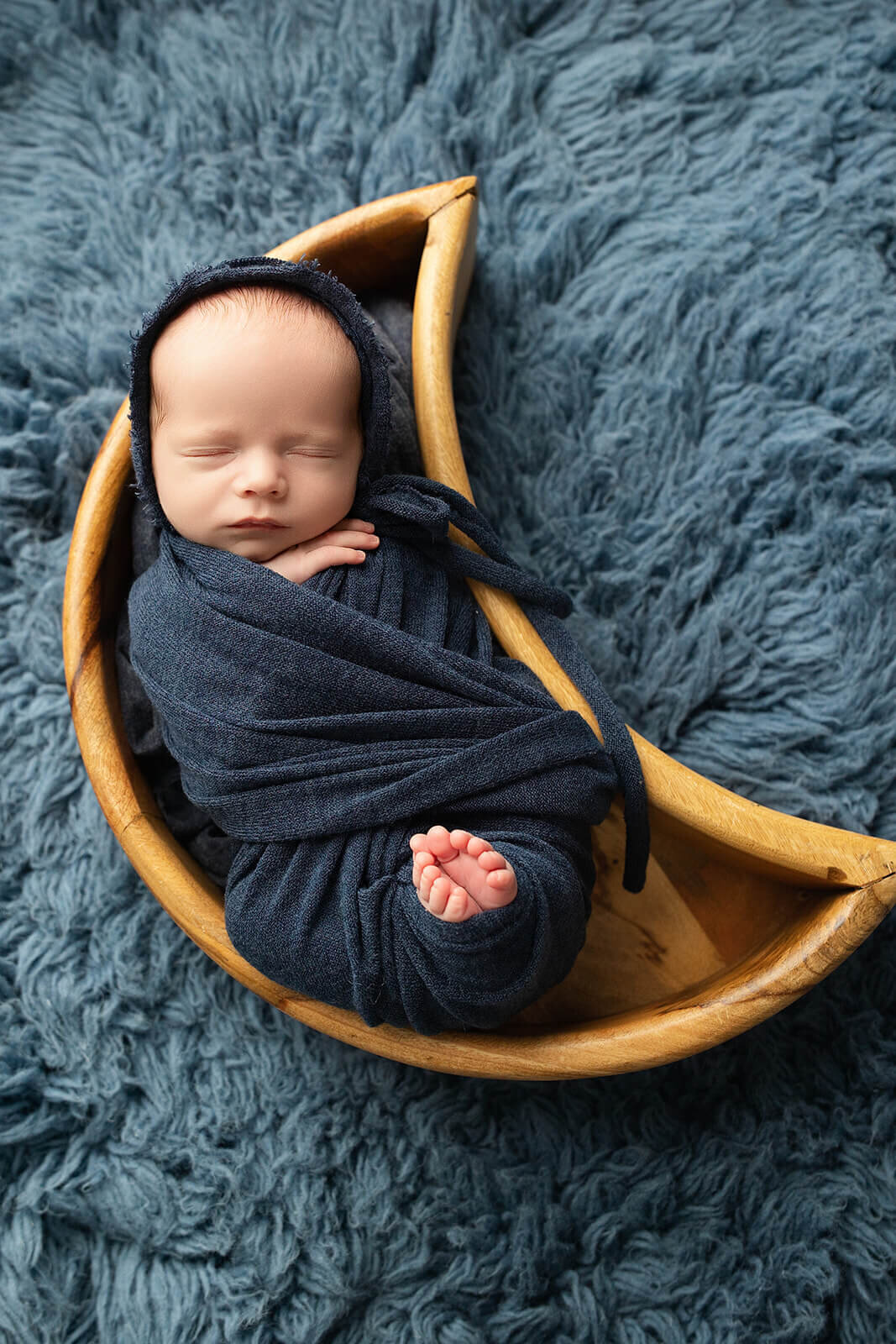 newborn baby boy wrapped with his toes peeking out in a navy blue wrap and matching bonnet posed in a wooden moon prop for york pa newborn photos 
