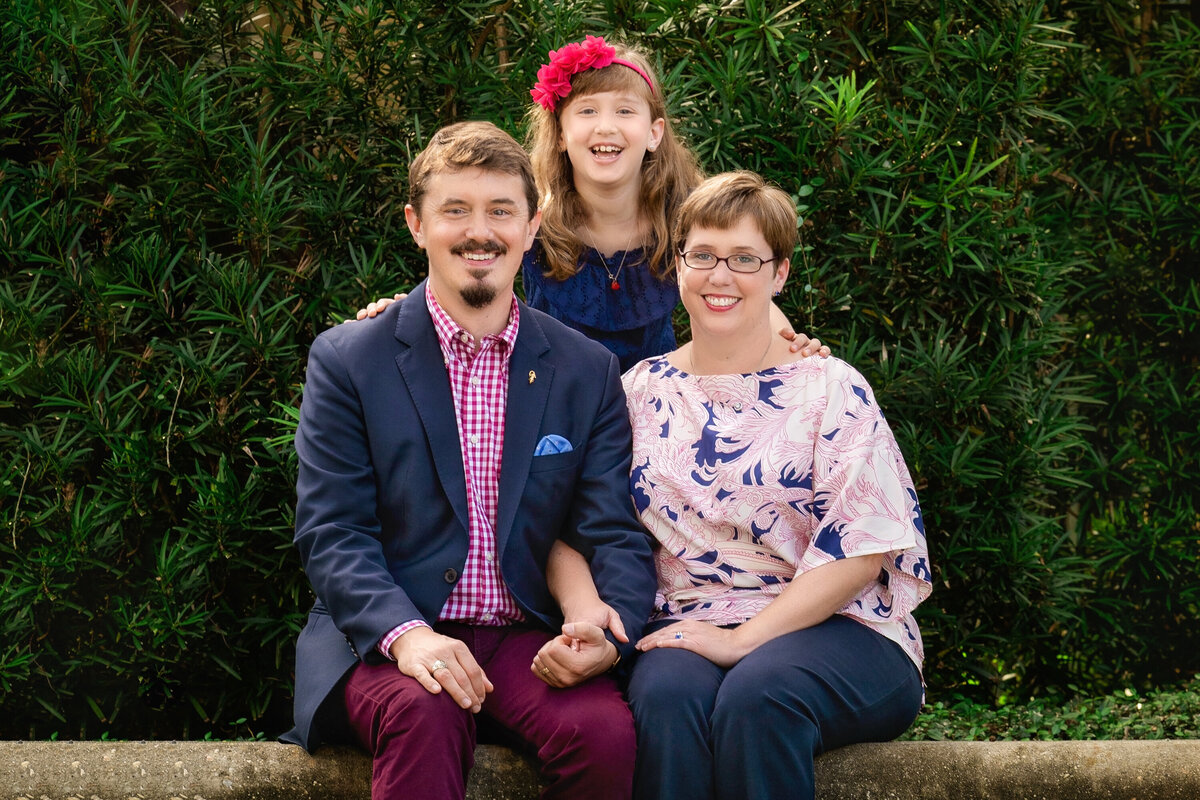 Mom, dad, and daughter are sitting on a bench at the Botanical Gardens in City Park.  Dad is wearing burgundy pants and a navy blazer.  Mom is wearing navy pants and a floral blouse.  Girl is wearing a navy dress and red floral headband.  They are all smiling at the camera.