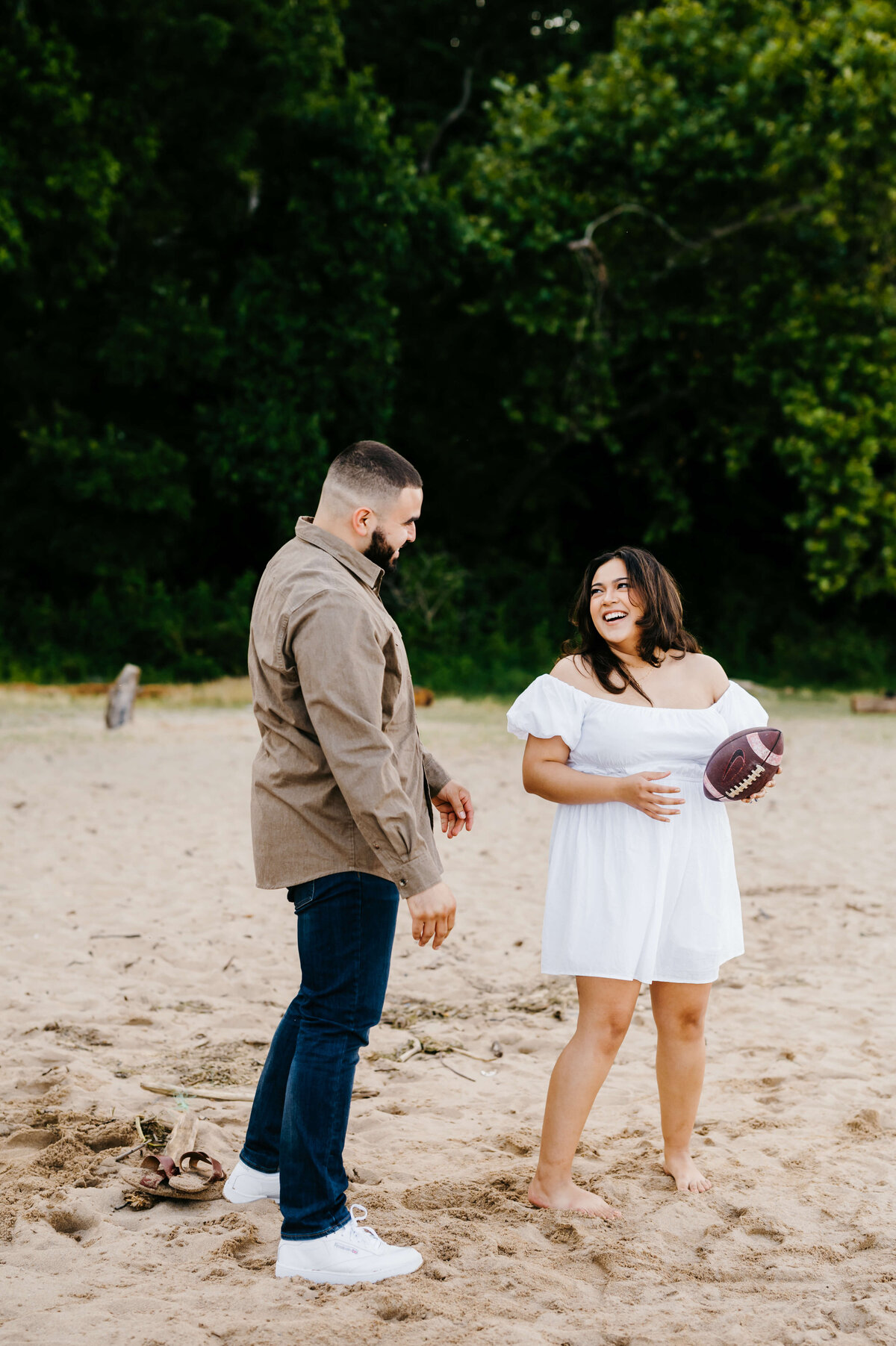 woman holding a football and laughing as she wears a white dress for engagement photos as a man in jeans and a brown shirt smiles at him on a lake beach with a lush wooded area in the distance