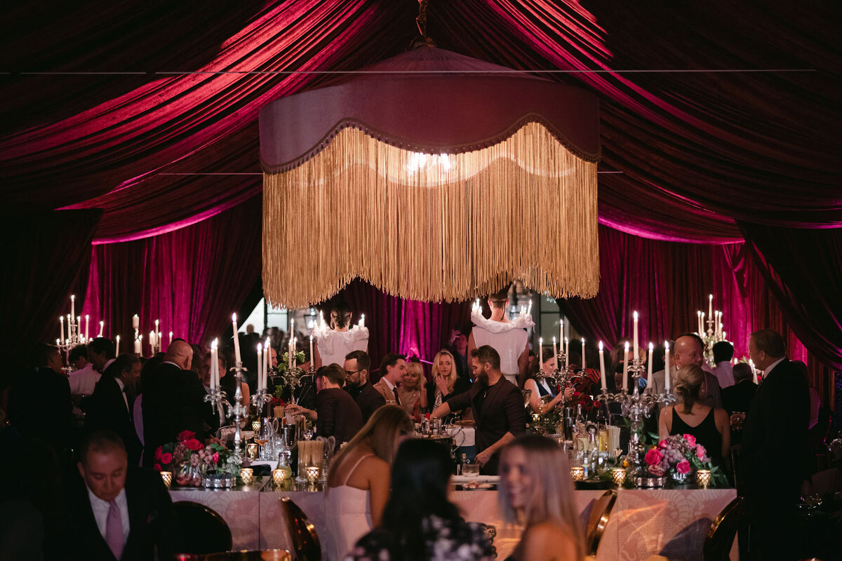 The photo was taken at a lavish indoor event in Virginia, a tented wedding captured beautifully by photographer Elisabetta Marzetti. Videographers Clayton Films also captured the stunning moments of the event. The attendees are seated at round tables under a grand, burgundy and gold draped canopy, surrounded by chandeliers, tall candles, and lavish floral arrangements. Guests can be seen engaged in conversation, truly relishing the elegant and sophisticated ambiance of the event.