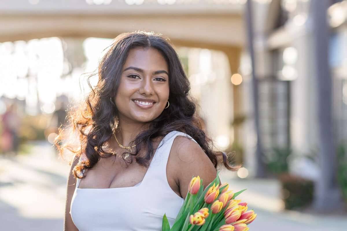 A woman with long wavy hair smiles while holding a bouquet of tulips. She is wearing a sleeveless white dress and is standing outdoors with blurred buildings and archways in the background.