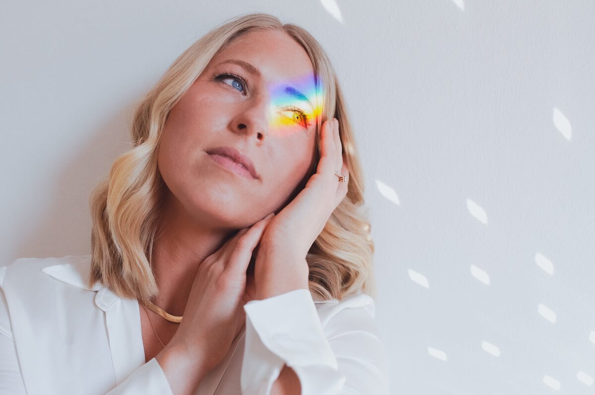 Woman with short, blonde, wavy hair softly smiles resting her head on her hand with reflected rainbow and disco ball shine on her face