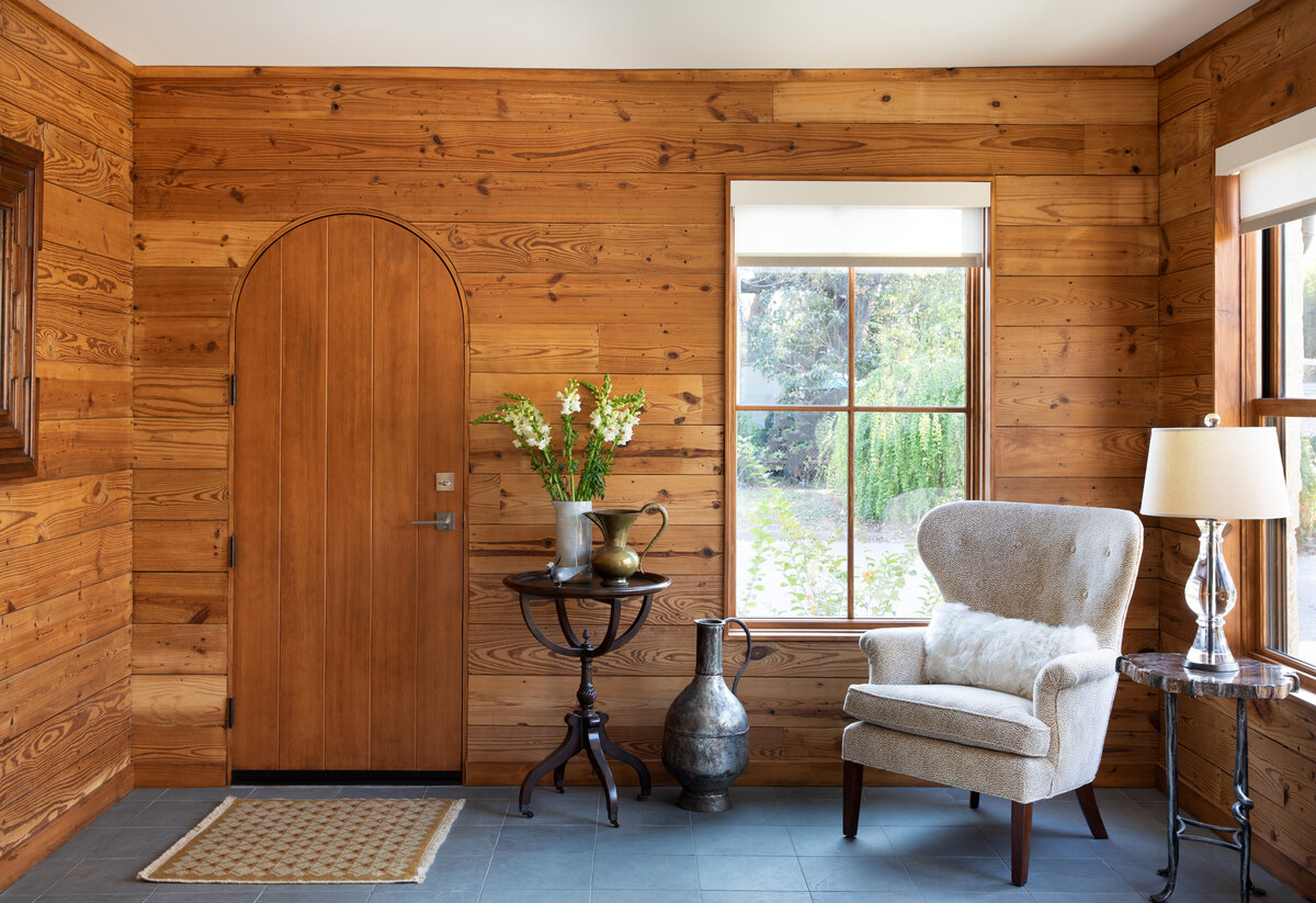 Dark warm wood tones cover the wall of this entryway. There is a rounded door and a large thin framed window. A boucle chair and old world side table are decor.