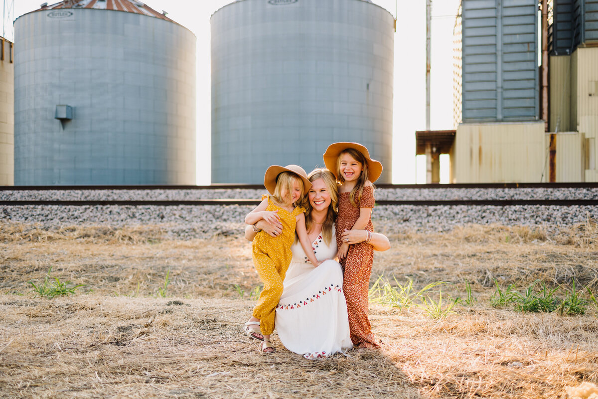 Family photo of mom with her two daughters on the railway, behind are two light blue water tanks. They are kneeling and hugging, the woman has a long white dress and the girls have orange and yellow dress with brown hat