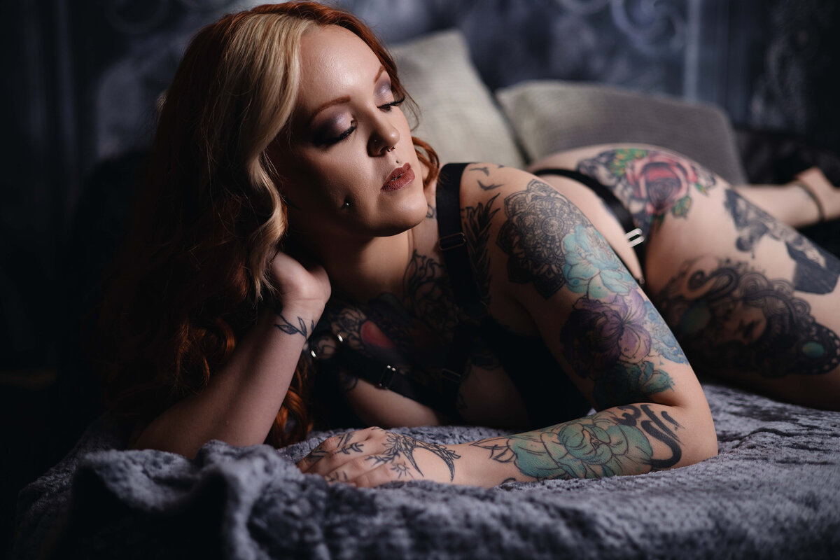 A person with long, wavy hair and numerous tattoos lies on their side on a bed with a gray blanket. They are wearing black lingerie and their eyes are closed, giving a serene expression.