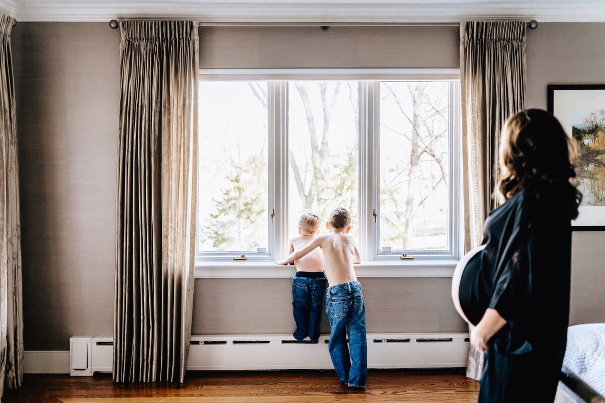 A pregnant woman stands in a bedroom, watching her two young sons as they stand on a windowsill, looking outside.