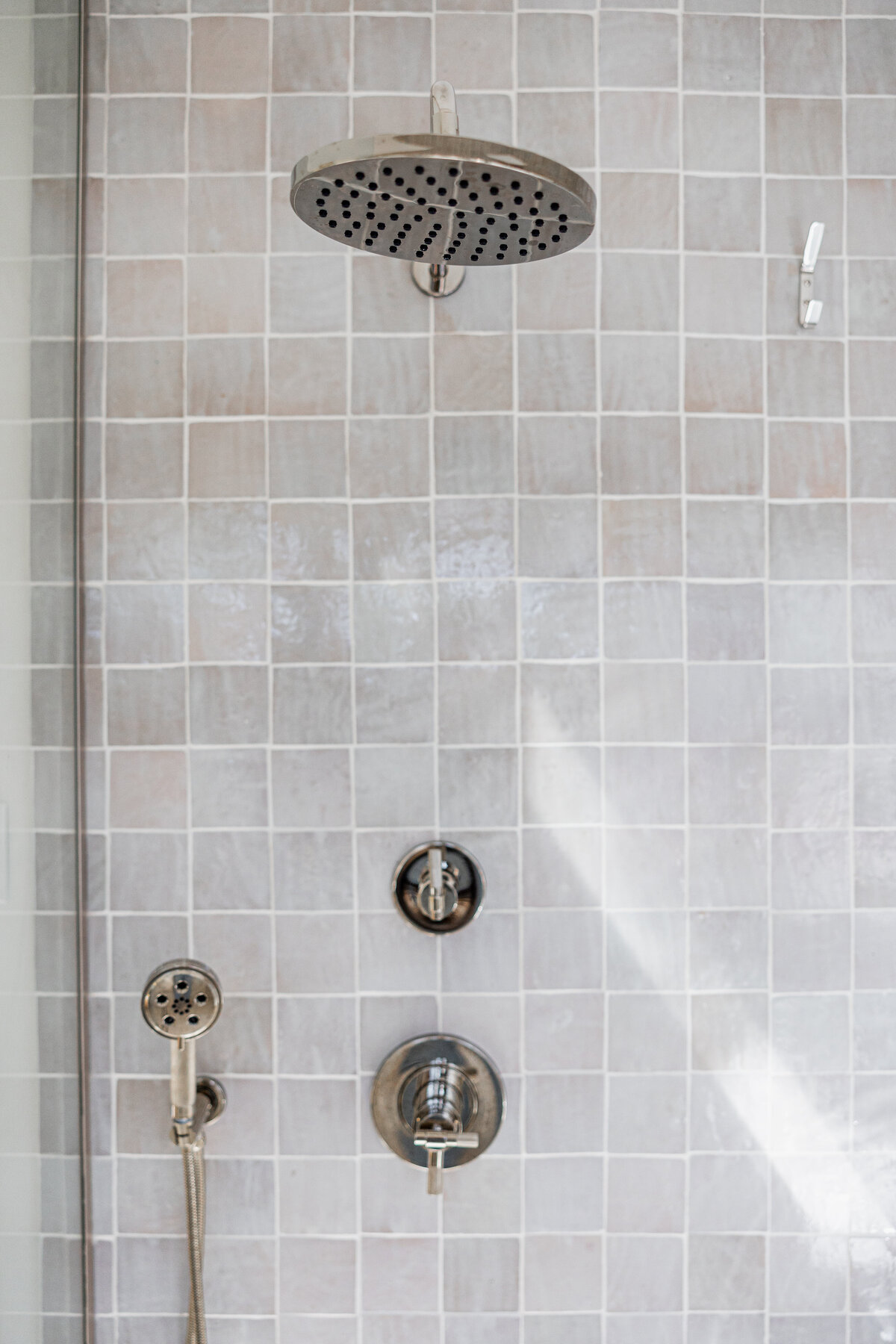 Close-up view of a modern bathroom shower featuring a large rainfall showerhead, handheld shower wand, and sleek silver fixtures against a backdrop of light grey tiled walls