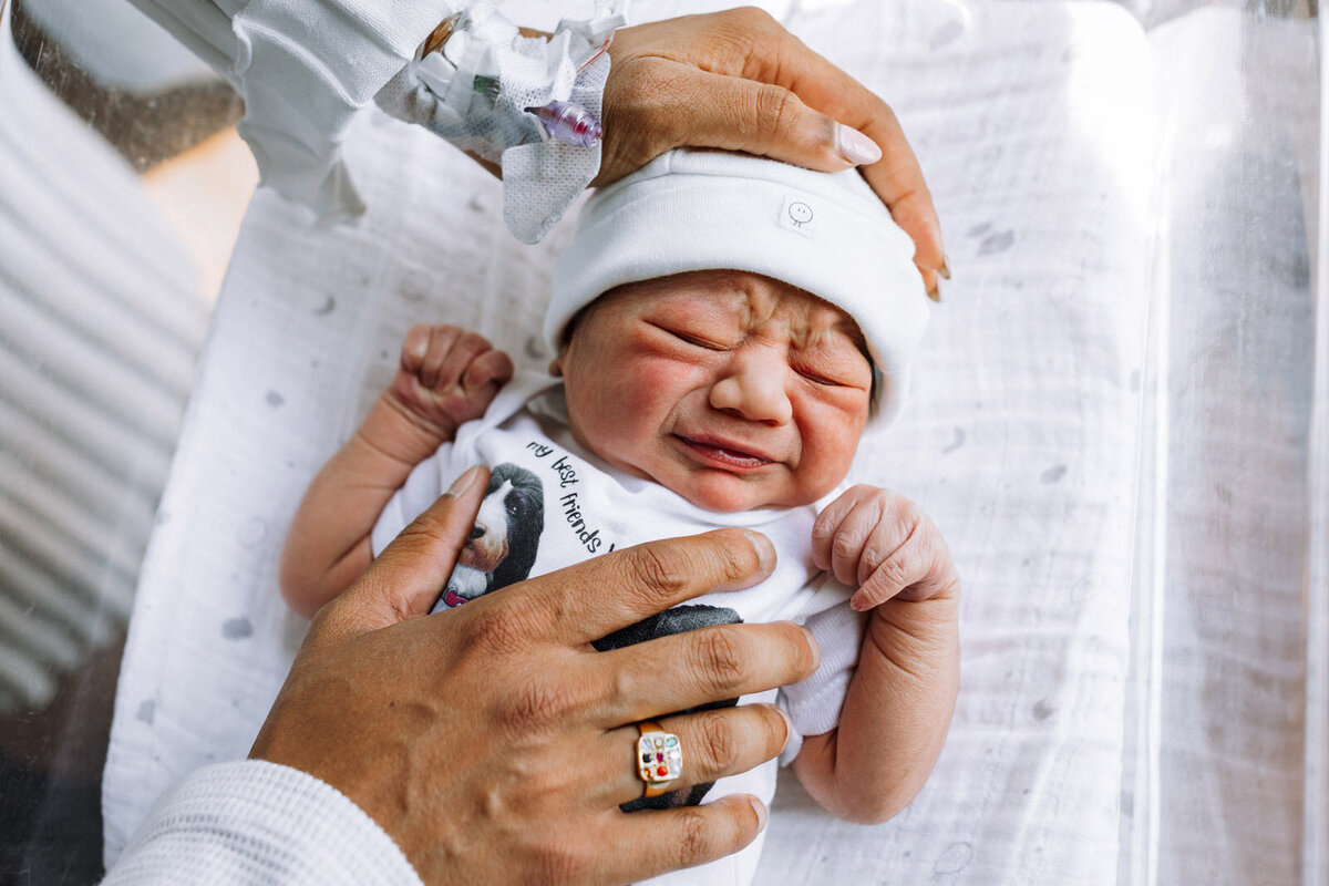 mom and dad's hands on crying newborn in hospital bassinet