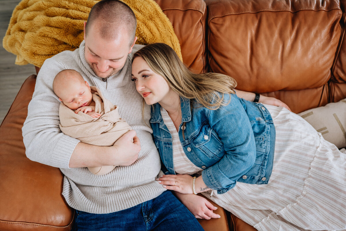 A family of three cuddles together on a leather couch, with the father cradling a swaddled newborn while the mother rests her head on his shoulder, all smiling peacefully.