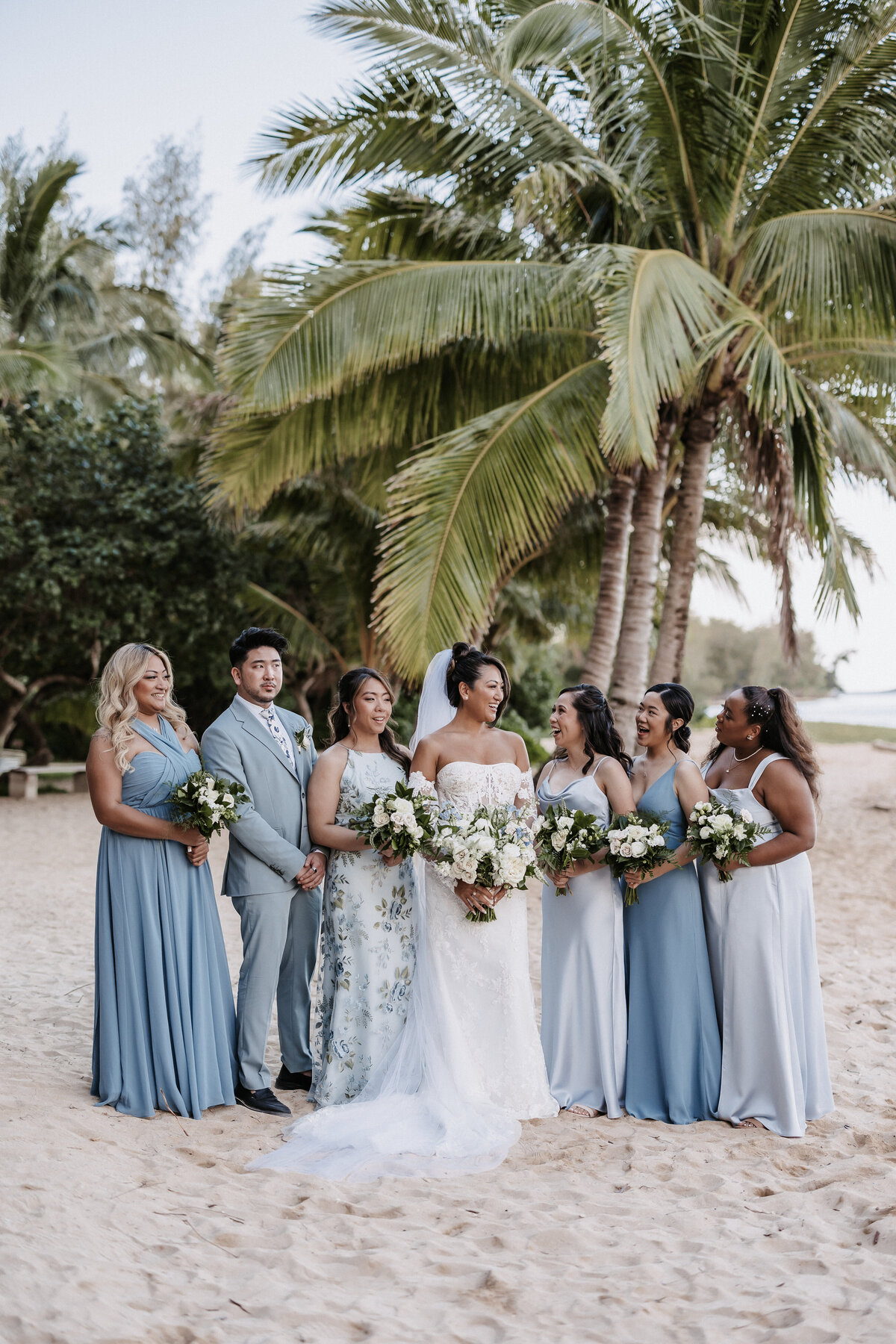 Bridesmaid and bridesman portraits wearing shades of blue holding bouquets on the beach with palm trees behind