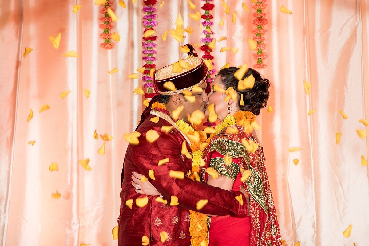 Hindu Groom and Bride under peach mandap  wearing red kissing as yellow rose petals fall from the sky . Hanging garlands of orange and magenta flowers hang behind them.