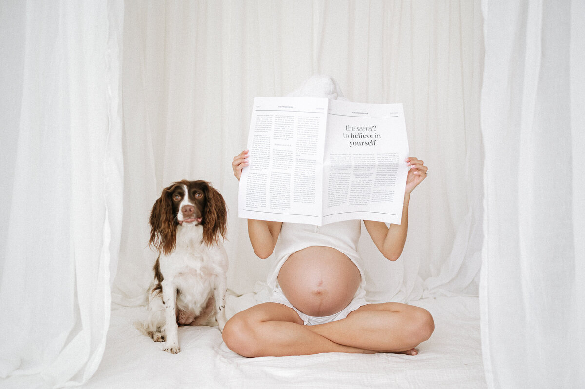 Pregnant woman sits reading her paper next to a dog during a maternity studio session