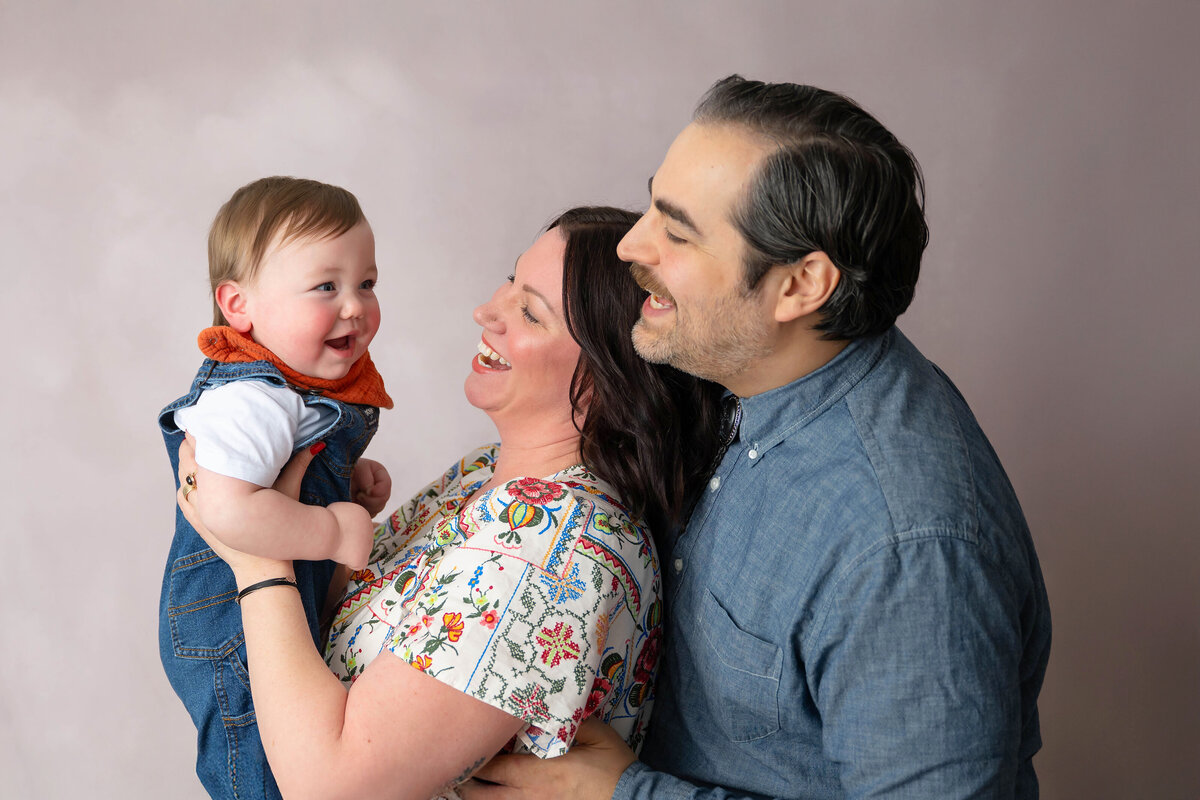 Mother holding baby while child looks at dad who is hugging mom from behind. 
