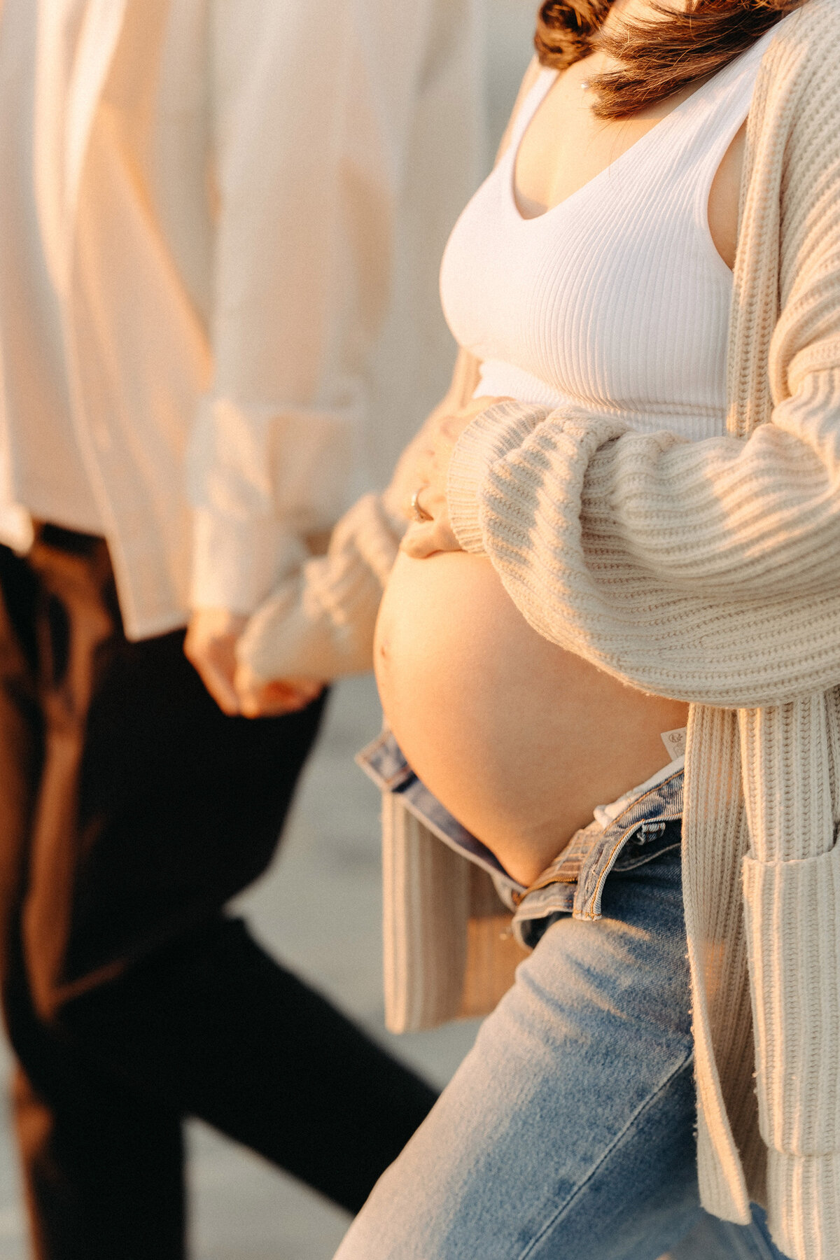 Close-up maternity photo of a mother in jeans and a cozy sweater, holding her baby bump while walking hand in hand with her partner at Coronado Beach. Captured by San Diego Family Photographer Yuka Schad.