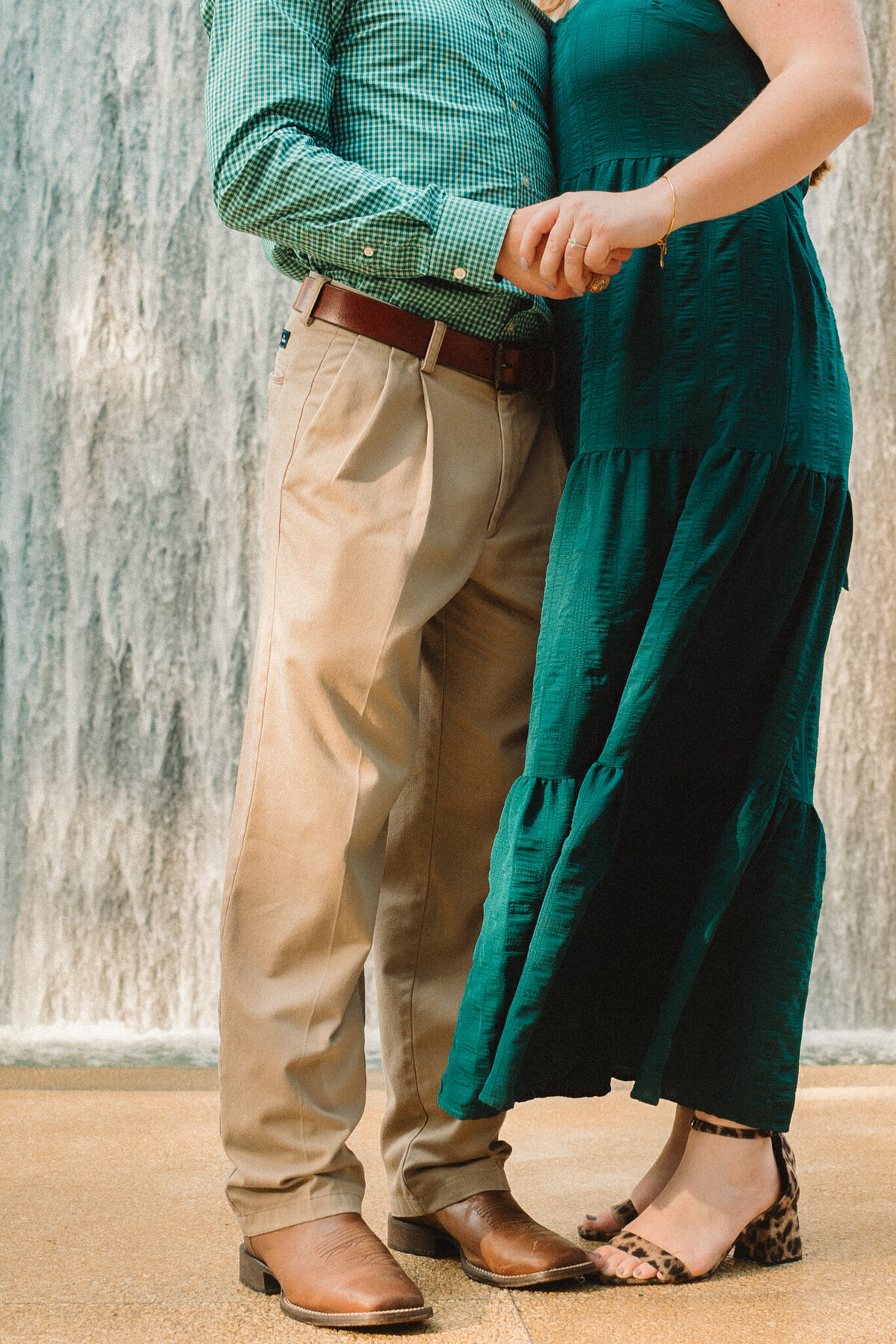 Close-up of a couple standing together during a proposal at the Gerald D. Hines Waterwall Park in Houston, Texas. The man wears tan pants and a green shirt, while the woman wears a flowing green dress against the iconic waterfall backdrop.