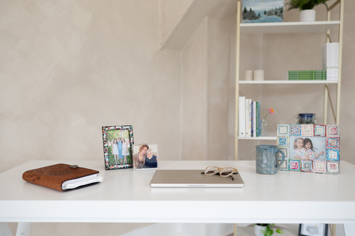 A white desk featuring a laptop, an open book, a photo frame, and a personal photo, creating a cozy workspace for a nutrition therapist.