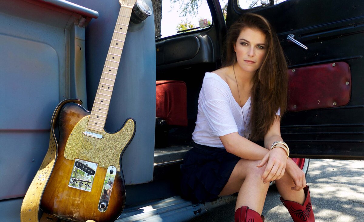 Female musician portrait Hayley wearing white shirt blue skirt red cowboy boots  sitting against vintage grey truck with electric guitar beside