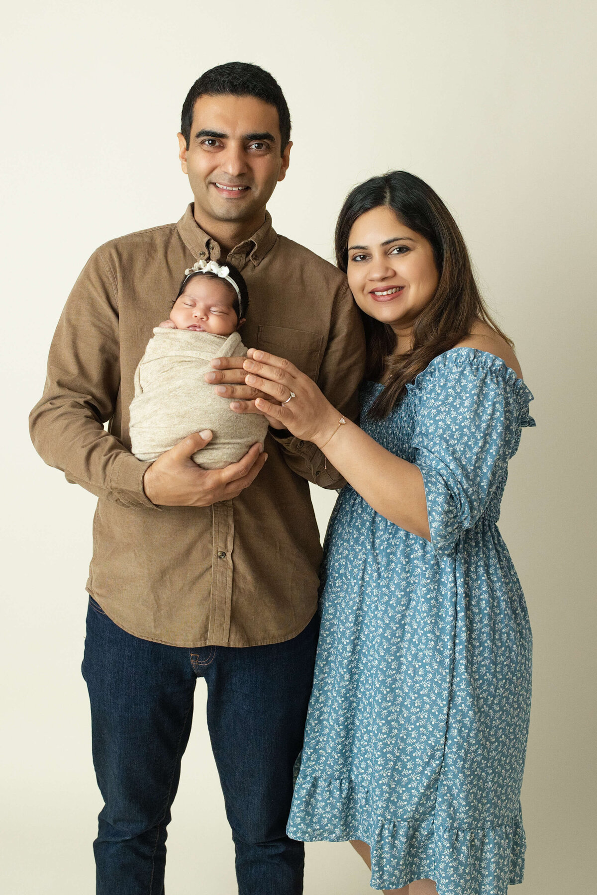 mom and dad holding their baby girl dad is in a tan shirt and mom in a blue dress on a light colored background posed for family photo