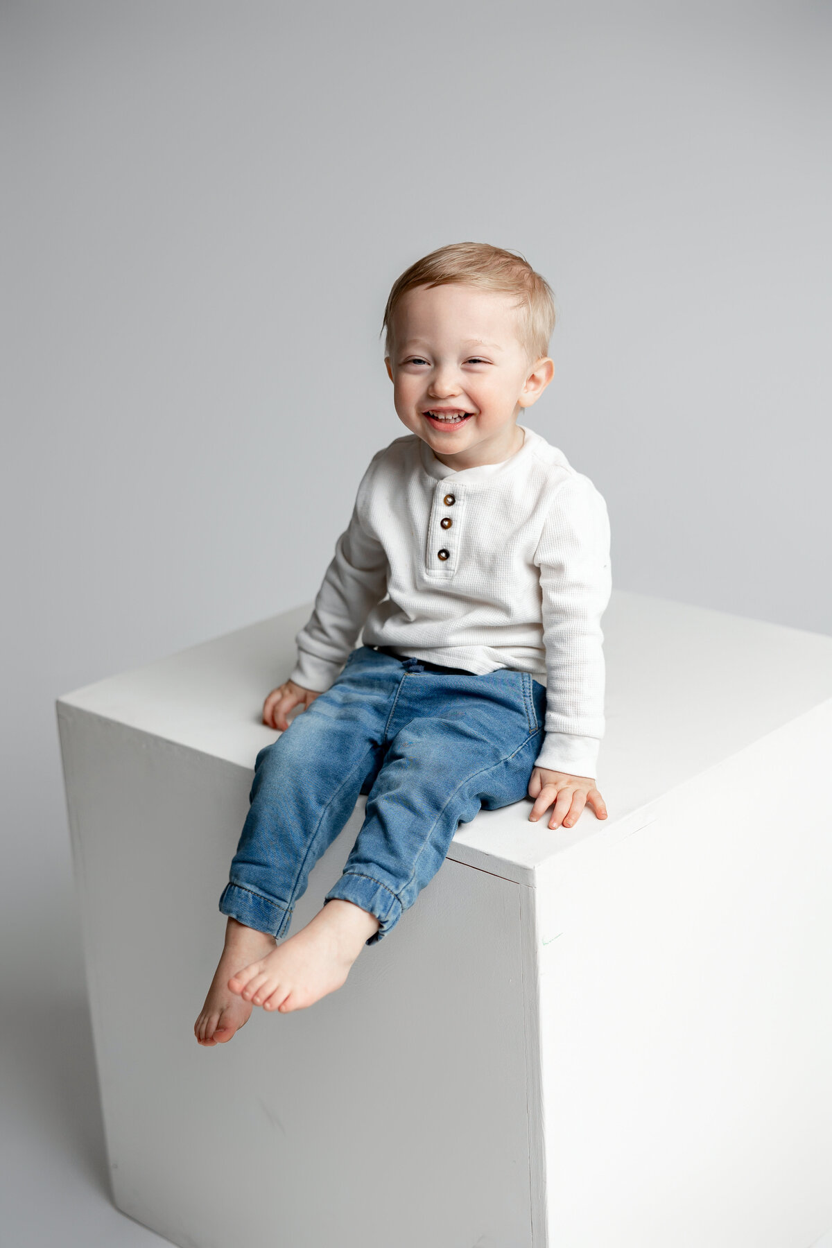 Toddler boy sitting on cube in jeans and light sweater, smiling in studio. Photographed by Yucaipa family photographer Kaitlyn Dawn Photography.