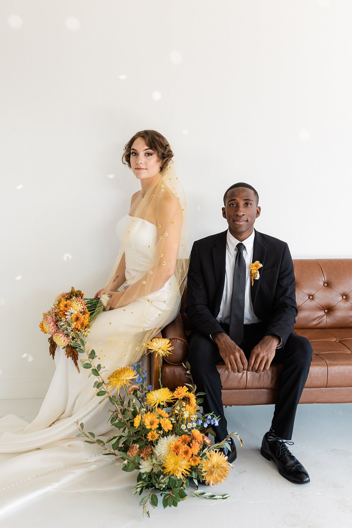 An editorial portrait of a bride and groom posing together on a leather couch on their wedding day in Dallas, Texas. The bride is on the left and is wearing an elegant, long, white dress with a golden veil and is holding a bouquet. The groom is on the right and is wearing a black suit with a boutonniere. A large floral arrangement rests on the ground between them.