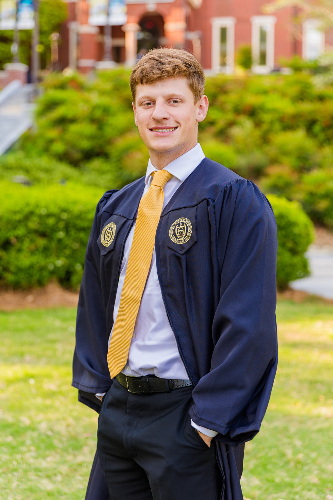 Senior boy standing on the college campus hands in pockets looking sideways at the camera wearing graduation gown during photo session with Atlanta photographer Laure Photography