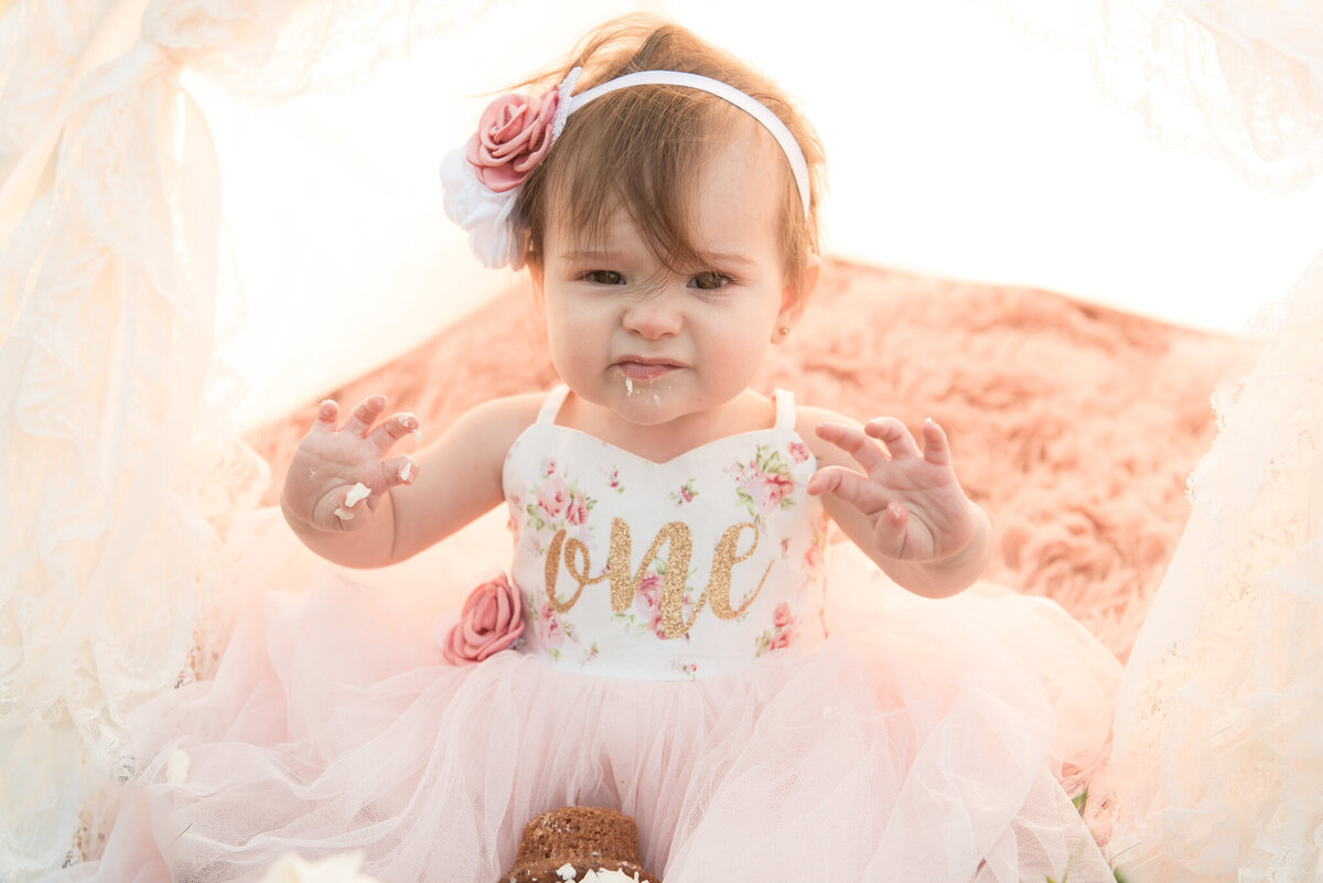 One year old girl looking at camera in lace tent with cake on her hands