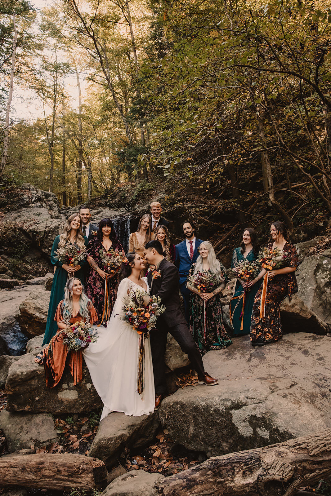 Newlyweds leaning in to kiss with their wedding parties behind them as they stand on rocks 