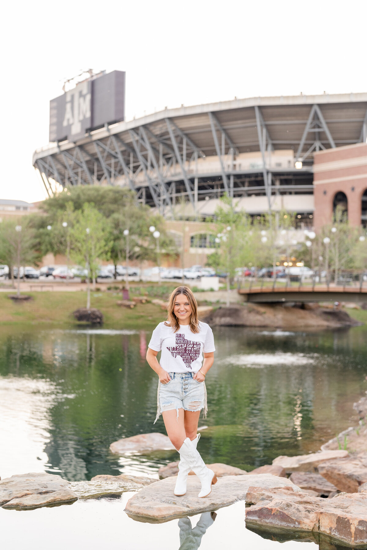 Texas A&M senior girl standing on rocks with hands in jean shorts pocket and smiling while wearing Aggie shirt in Aggie Park with Kyle Field in the background