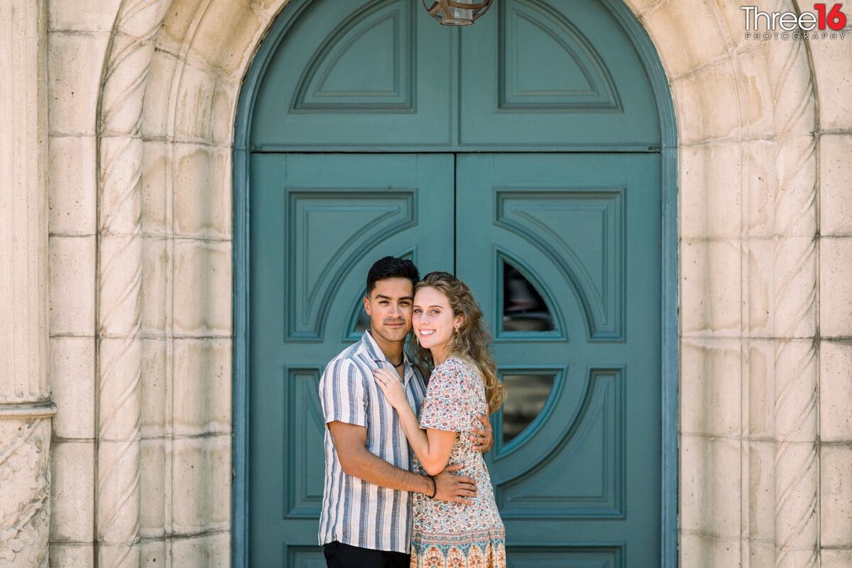 Engaged couple pose together cheek to cheek in front of the Claremont Train Station green door entrance during engagement photo session