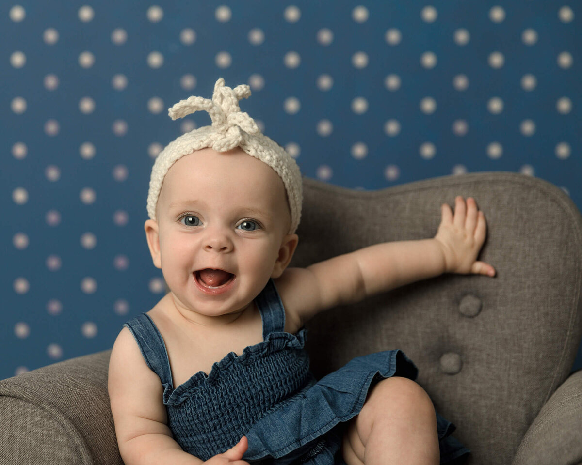 Baby girl with white headband sitting in a grey chair in front of blue and while polka dot background.