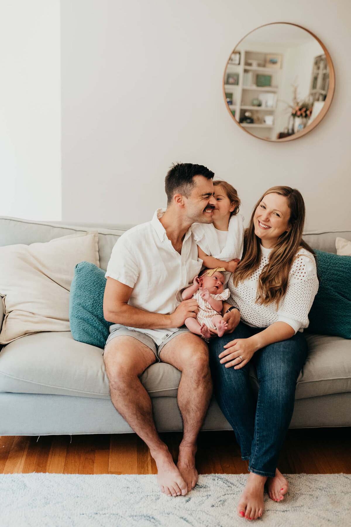A family sit on their couch together holding their newest baby, the mother smiles at the camera while the toddler daughter kisses her father on the cheek
