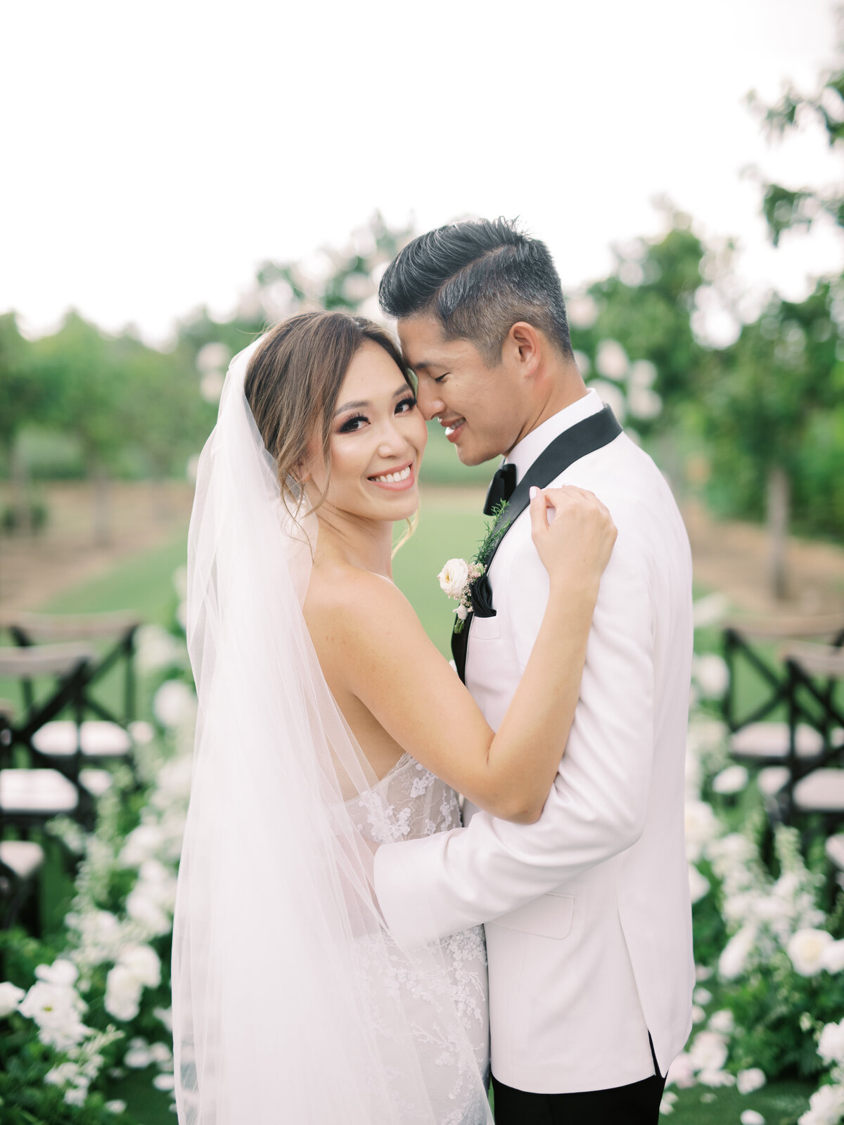 Kohnur Romantic couple close-up with bride in soft glam makeup and loose waves wearing a strap gown and veil, embracing on an orchard wedding aisle