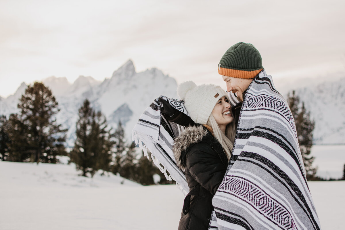 winter engagement session in the Grand Tetons man araps up his fiance in a boho blanket as they stand in front of a mountain range