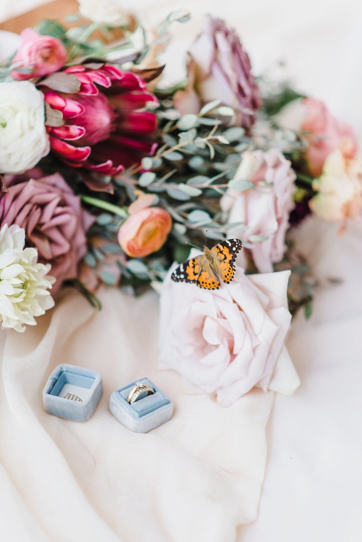 A live monarch butterfly  sits on a rose from a bouquet laying sideways on soft material with a dusty blue ring box and diamond ring laying beside it at a wedding  in Ottawa Ontario
