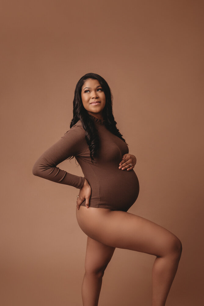 Maternity session with Atlanta, Georgia maternity photographer pregnant woman holding baby bump wearing dark brown body suit on tan backdrop