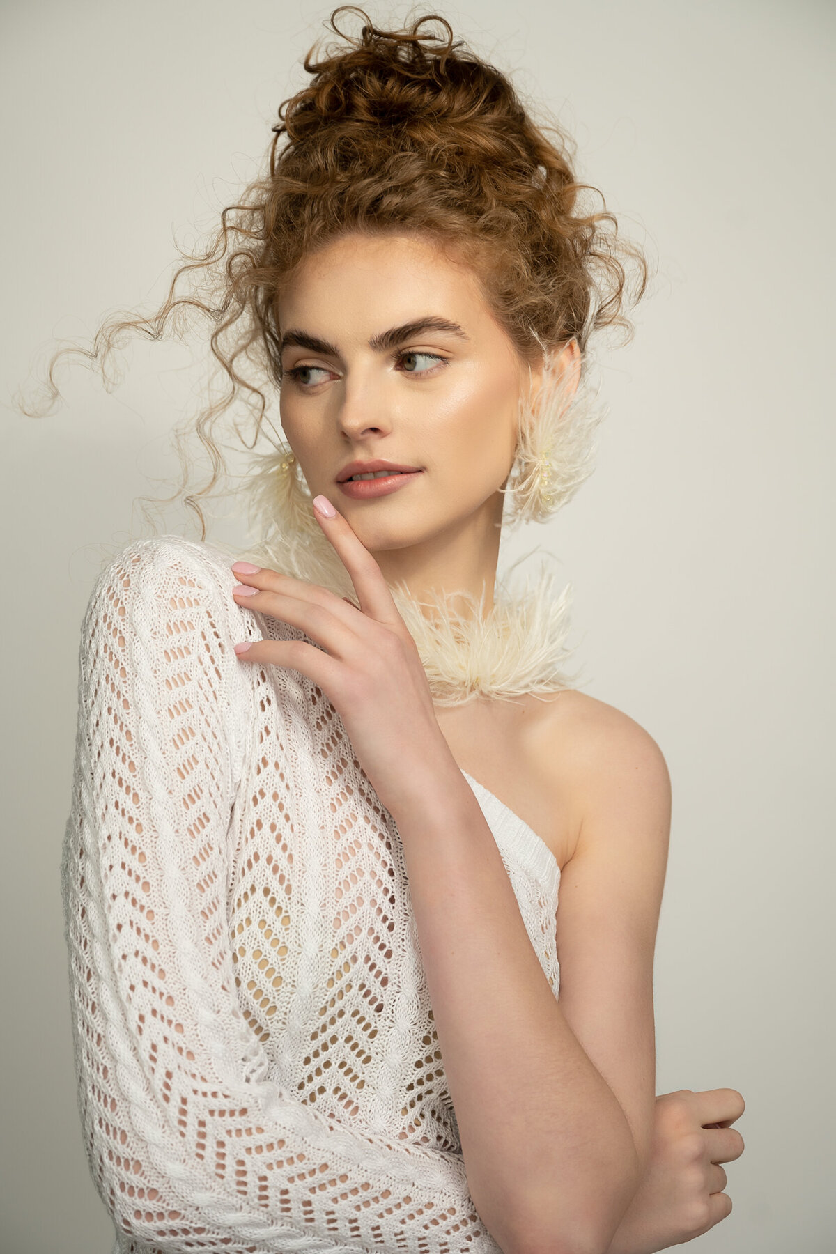 A woman wearing a one sleeve white gown and a feather necklace. She is touching her lipt with her hand