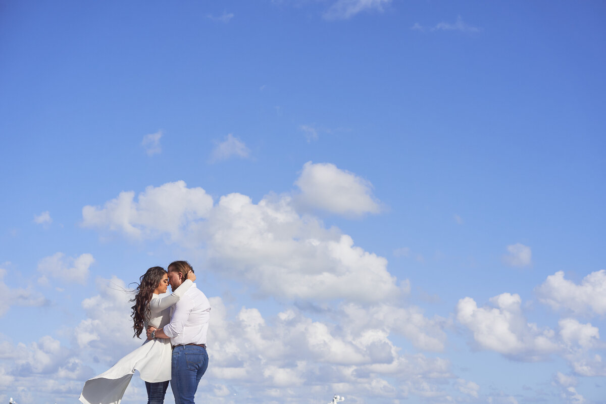 Couple embracing under a bright blue sky filled with fluffy white clouds, capturing a romantic moment on a sunny day in Miami.