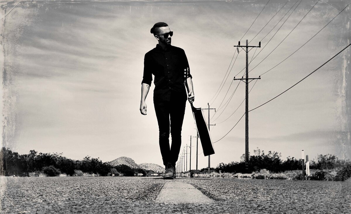 Male musician photo black and white Michael Bernard Fitzgerald wearing black outfit caring guitar walking down center road  desert background