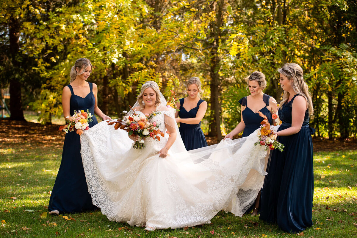 bridesmaids wearing long navy gowns hold a bride's wedding dress off the ground as she walks to her Ottawa wedding at Strathmere