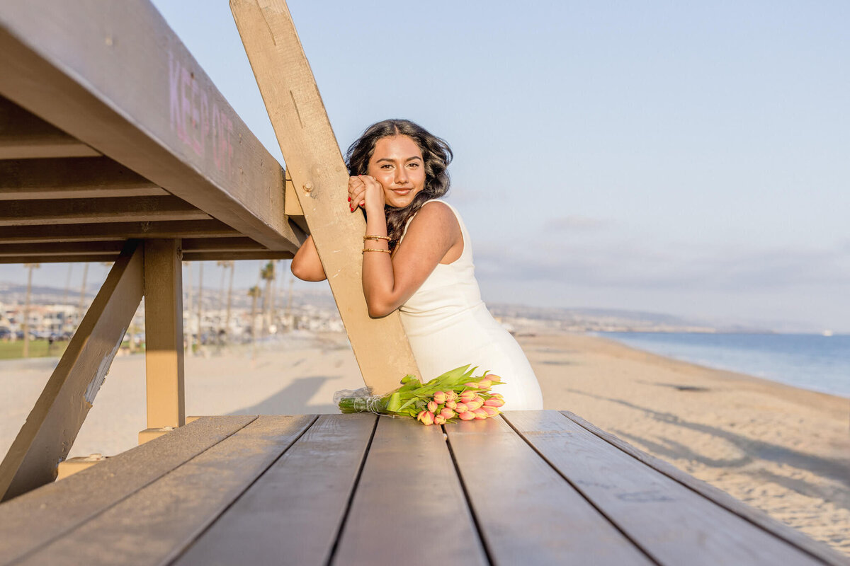 A woman in a white dress leans against a lifeguard tower on a sunny beach. She gazes at the camera, with a bouquet of tulips resting on a wooden table in the foreground. The ocean and clear sky form the backdrop.