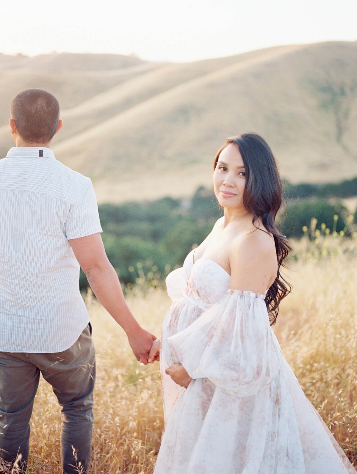 Maternity photoshoot in San Francisco with a pregnant woman and expecting dad standing in a field against hills.