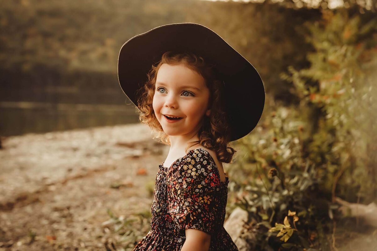 A girl wearing a floral dress and black hat looks off into the distance while smiling in Rochester ny
