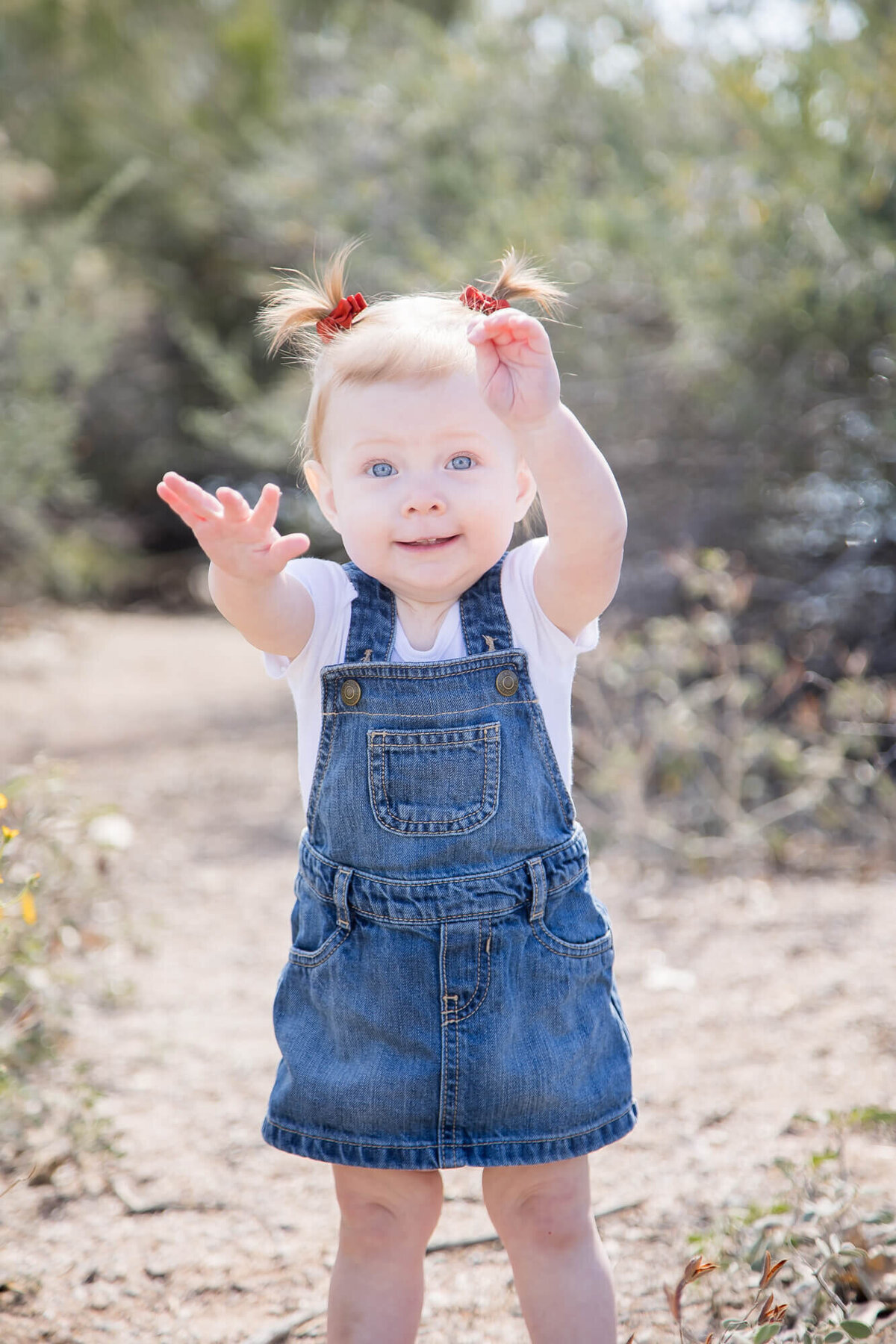 Toddler raising her arms while wearing a denim dress and pigtails on desert park path