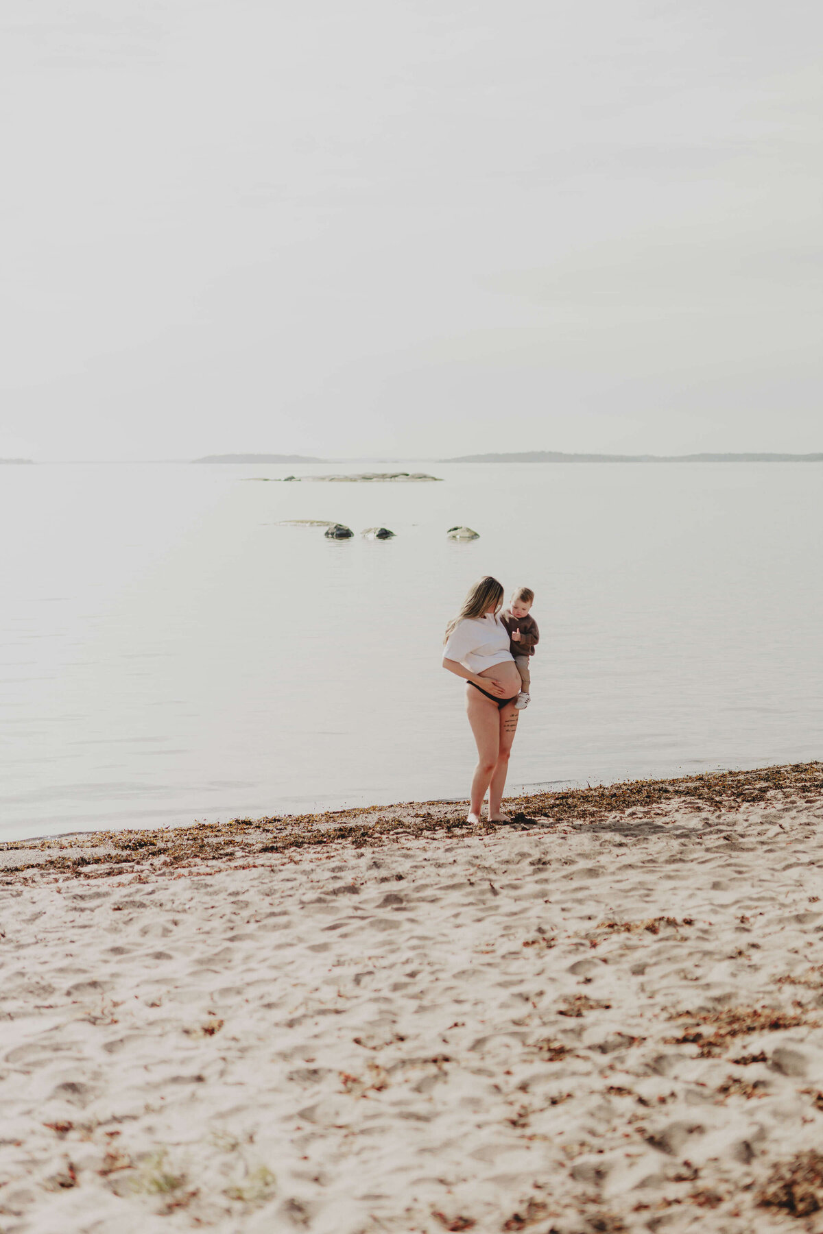 En gravidbild på en gravid kvinna hållandes sin son längre bort på en strand under en gravidfotografering i Stockholm