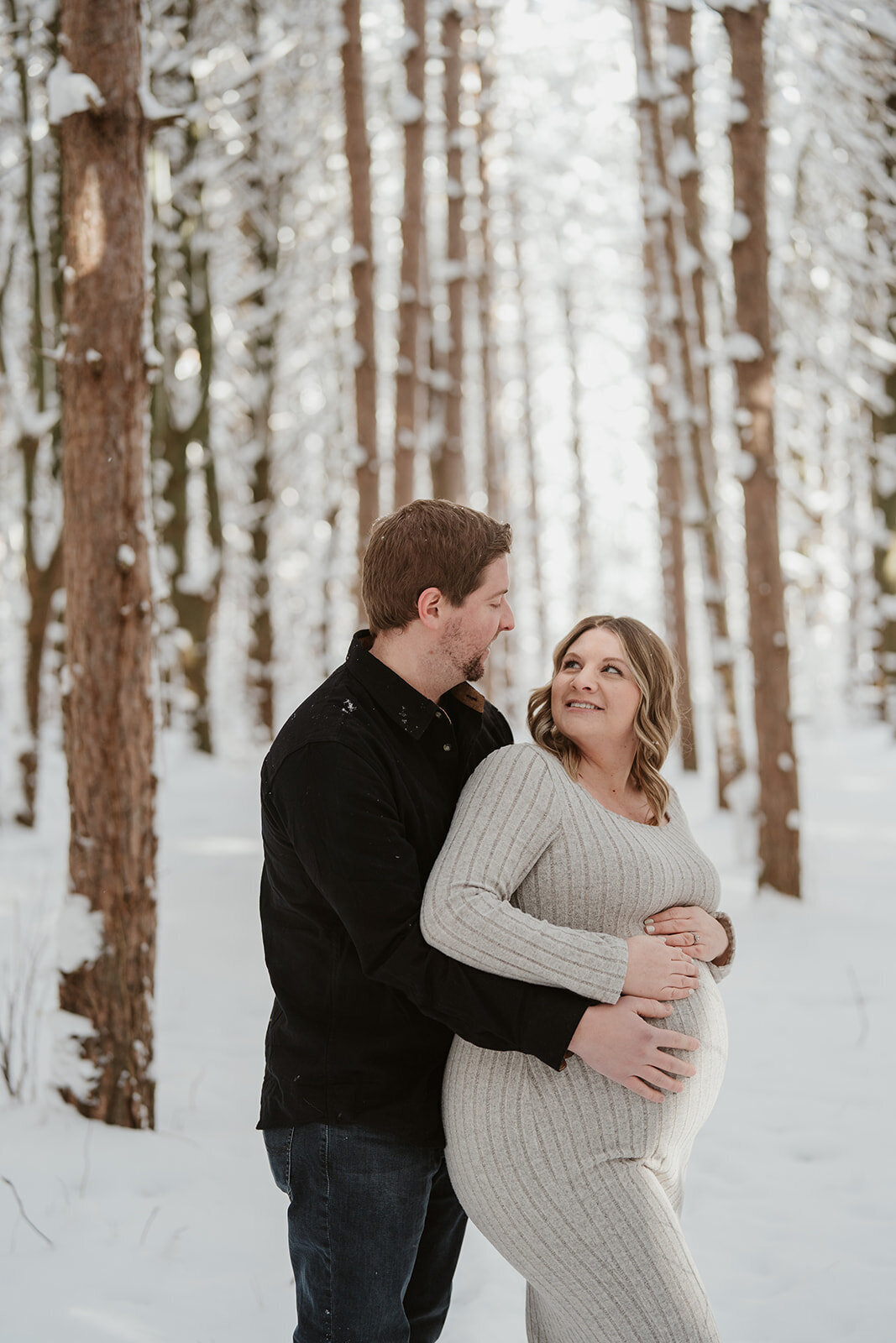Pregnant woman posing in front of snow-covered pine trees at Al Sabo Preserve during her winter maternity session in Kalamazoo.