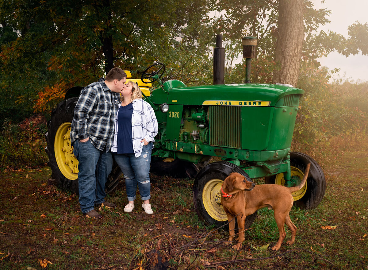 Couple kissing by green tractor with dog nearby in rustic setting