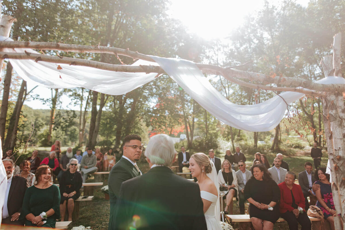 An outdoor wedding ceremony under soft sunlight, with the couple exchanging vows surrounded by family and friends.